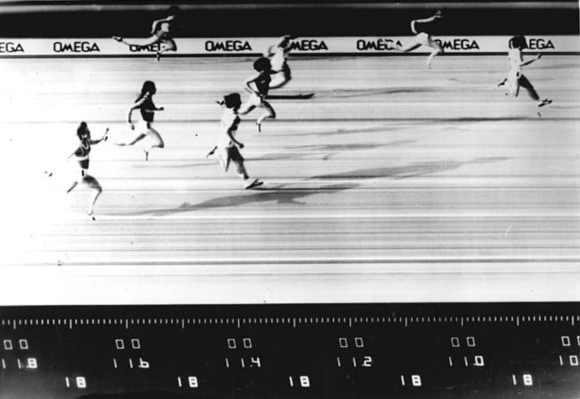A photograph of a race finish line captured at the moment two athletes cross, with a timing display.