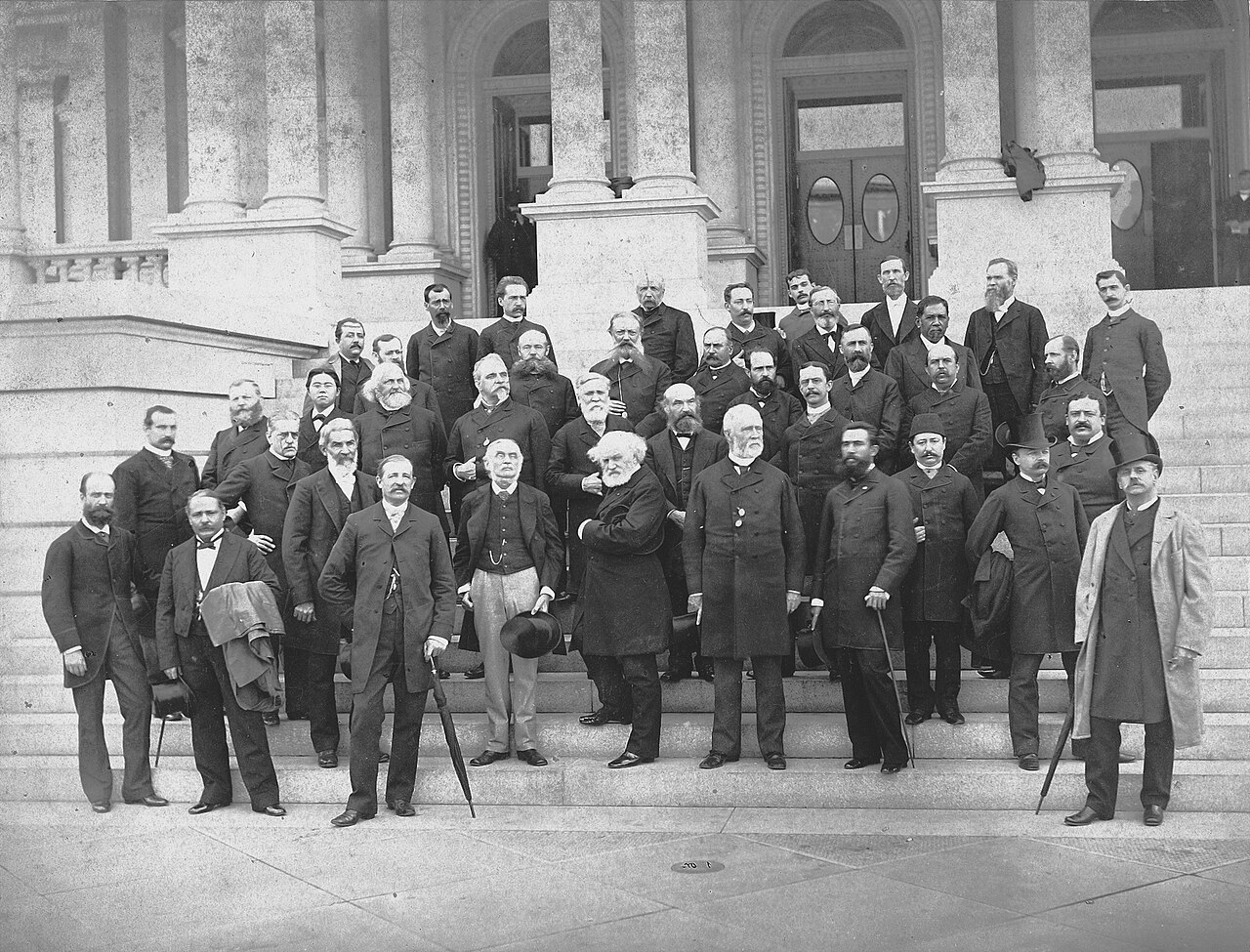 Black-and-white group photograph of formally dressed nineteenth-century delegates arranged in rows on stone steps before a columned building.