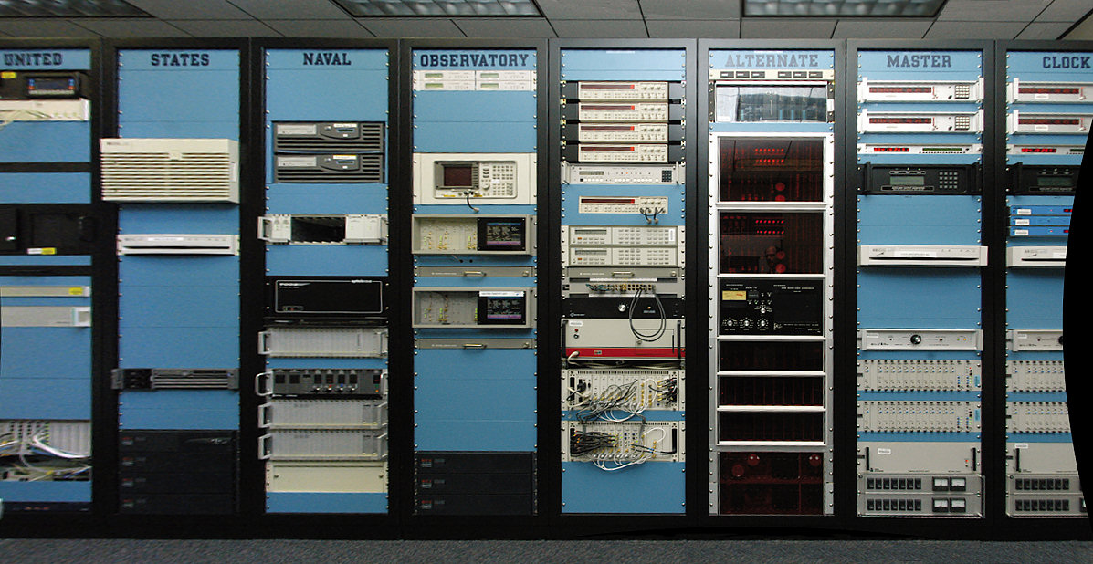A equipment room of tall rack cabinets stacked with electronic timekeeping instruments and cables, overhead fluorescent lighting, no people.